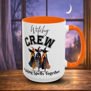 May include: A white ceramic mug with an orange interior and handle. The front features the text "Witchy CREW Making Spells Together" in black lettering above an illustration of three figures wearing pointed hats and long dresses.