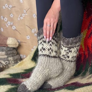 May include: A pair of thick, fuzzy, hand-knit wool socks featuring a neutral color palette of beige, cream, and dark brown. The socks display a geometric pattern around the calf and solid dark brown toes, crafted with a visible, chunky knit texture.