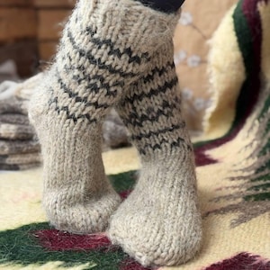 May include: A pair of thick, hand-knitted wool socks in a natural beige color. The socks feature a ribbed cuff and a decorative pattern of dark charcoal horizontal zigzag stripes around the ankle area. The texture is fuzzy and chunky.