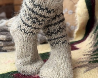 May include: A pair of thick, hand-knitted wool socks in a natural beige color. The socks feature a ribbed cuff and a decorative pattern of dark charcoal horizontal zigzag stripes around the ankle area. The texture is fuzzy and chunky.