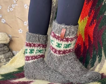 Puede incluir: Un par de calcetines de lana gruesa tejidos a mano en color gris, con puño acanalado y una banda decorativa con patrones geométricos en granate, crema y verde. Una pequeña etiqueta de cuero marrón está unida al puño superior derecho.