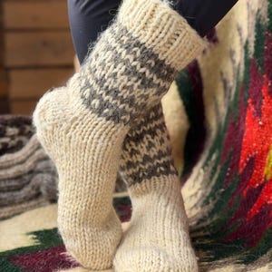 May include: A pair of thick, hand-knitted wool socks featuring a cream-colored base with a contrasting dark gray patterned band around the upper calf. The socks have a ribbed cuff at the top and a chunky, textured knit construction throughout.