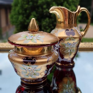 Puede incluir: Un tarro de cristal rojo rubí con borde dorado, con tapa y una jarra a juego. Ambos están adornados con diseños florales en blanco, azul y dorado. El tarro y la jarra se reflejan en una superficie espejada.