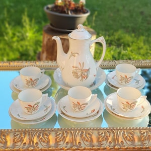 Puede incluir: Juego de té de porcelana blanca con diseños florales, que incluye una tetera, seis tazas y platillos. El juego se exhibe sobre una superficie espejada con un marco dorado ornamentado, sobre un fondo verde borroso.