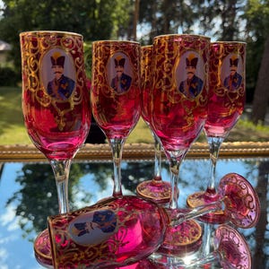 Puede incluir: Un juego de seis copas de flauta de cristal rojo rubí con filigrana dorada y un retrato ovalado central de un hombre con uniforme militar. Las copas tienen tallos y bases transparentes, con una copa colocada horizontalmente sobre las demás.