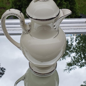 May include: Cream-colored porcelain teapot with silver trim and a cherub figurine on the lid. The teapot has a rounded body, a curved handle, and a spout. The image is reflected on a mirrored surface.
