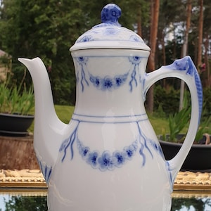 May include: White porcelain coffee pot with a blue floral design. The pot has a curved spout, a handle, and a lid with a blue knob. The floral design includes garlands and small flower clusters. The pot is sitting on a reflective surface.