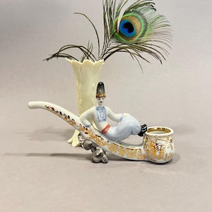 May include: A decorative porcelain pipe with a seated figure in traditional dress. The pipe is white with gold detailing and a small dog figurine. A peacock feather and a cream vase are in the background.