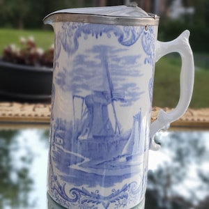 May include: A white and blue ceramic pitcher with a silver lid. The pitcher features a Dutch windmill scene. The handle is white and has a unique, decorative shape. The pitcher is reflected in a mirrored surface.