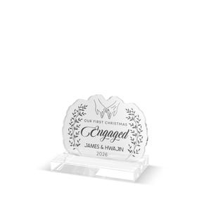 Puede incluir: Una placa de acrílico transparente con base rectangular que presenta un grabado de dos manos entrelazadas, ramas con hojas a los lados y el texto "OUR FIRST CHRISTMAS Engaged JAMES & HWAJIN 2026" centrado en la superficie frontal.