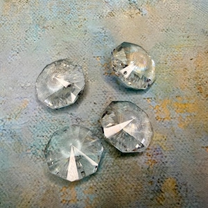 May include: Four clear, faceted crystal pendants with multiple sides, reflecting light. The crystals are arranged on a textured background with blue, gray, and gold tones. These decorative crystals could be used for jewelry or home decor.
