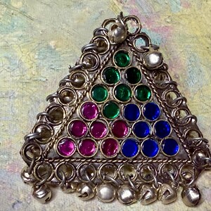 May include: Silver triangular pendant with a vibrant array of gemstones. The pendant showcases green, pink, and blue stones, and is embellished with small dangling bells. The design is intricate and detailed.