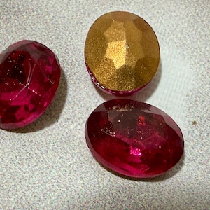 May include: Three gemstones are displayed on a light gray surface. Two are oval-shaped, faceted, and a deep red color. The third is round, with a gold faceted top and a red base. The gemstones are likely for jewelry making.