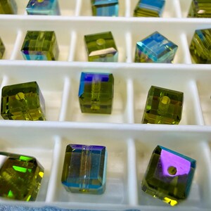 May include: Close-up of iridescent cube-shaped beads in a white tray. The beads are a mix of green, blue, and purple, with a hole drilled through the center. The beads are arranged in rows.