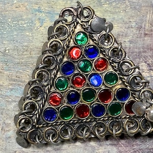 May include: A triangular pendant with a silver-toned metal frame. The frame is decorated with small circular accents and chains. The pendant is filled with a pattern of red, green, and blue glass stones. A small silver charm hangs from the bottom.