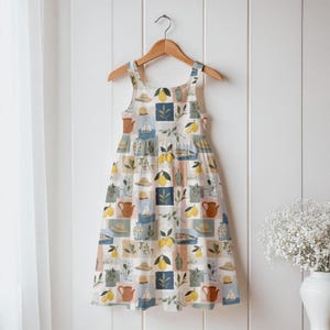May include: A sleeveless dress with a square neckline, hanging on a wooden hanger. The dress features a repeating pattern of lemons, hats, teapots, and sailboats in a patchwork design. The color palette includes white, blue, yellow, and brown.
