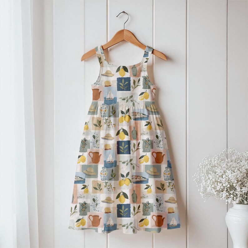 May include: A sleeveless dress with a square neckline, hanging on a wooden hanger. The dress features a repeating pattern of lemons, hats, teapots, and sailboats in a patchwork design. The color palette includes white, blue, yellow, and brown.