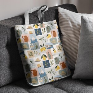 May include: A white tote bag with a repeating pattern of lemons, hats, sailboats, and teapots in a patchwork design. The bag has gray handles and is resting on a gray sofa next to striped and solid pillows.