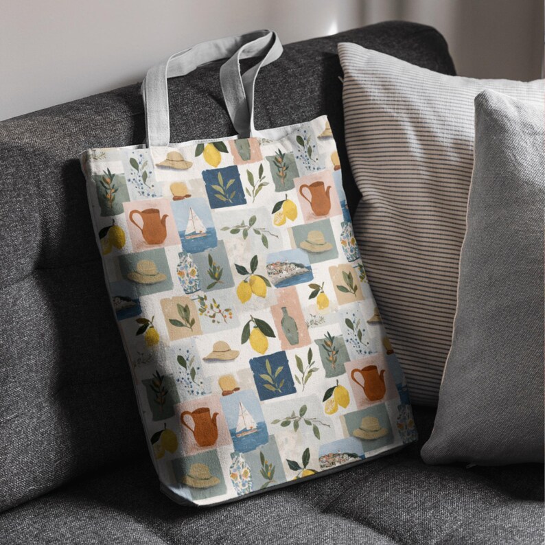 May include: A white tote bag with a repeating pattern of lemons, hats, sailboats, and teapots in a patchwork design. The bag has gray handles and is resting on a gray sofa next to striped and solid pillows.