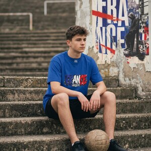 May include: A person wearing a blue t-shirt with the word "FRANCE" in red, white, and blue, sitting on concrete steps. A vintage soccer ball rests in front of the person. The background includes a weathered poster and stadium seating.