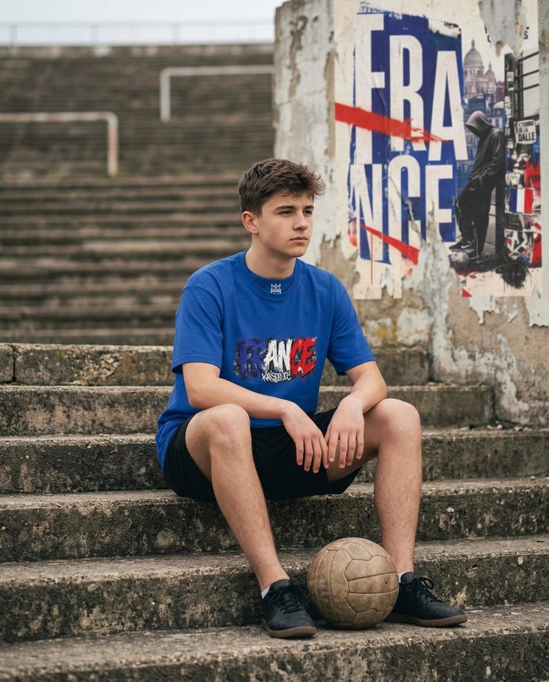 May include: A person wearing a blue t-shirt with the word "FRANCE" in red, white, and blue, sitting on concrete steps. A vintage soccer ball rests in front of the person. The background includes a weathered poster and stadium seating.