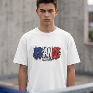 May include: White t-shirt with the word "FRANCE" in blue and red, with "Kaiser FC" below. The design has a distressed, paint-splatter effect. The shirt is worn by a person in front of a concrete background.