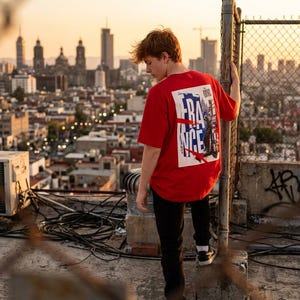 May include: A red t-shirt with a graphic on the back featuring the word "FRANCE" in white and blue, with a cityscape illustration. The person is wearing black pants and sneakers, standing on a rooftop with a city skyline in the background.