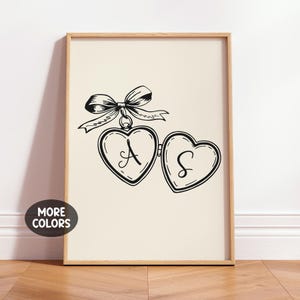 May include: A framed print featuring a black and white illustration of a heart-shaped locket with the letters "A" and "S" inside. A decorative bow is above the locket. The print is displayed on a cream-colored background.