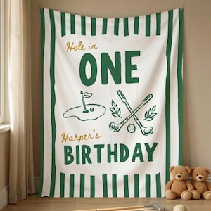 May include: A white and green striped fabric banner featuring the text Hole in ONE and Harper's BIRTHDAY in dark green and gold. The design includes a golf hole illustration, two crossed golf clubs, and leafy accents on a white background.