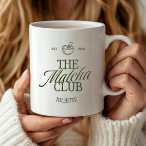 May include: A white ceramic mug featuring the text "THE Matcha CLUB" in a script font, with the name "Juliette" below. The mug also has a small graphic of a bowl and whisk, and the text "EST 2002". The mug is held by a person wearing a white sweater.