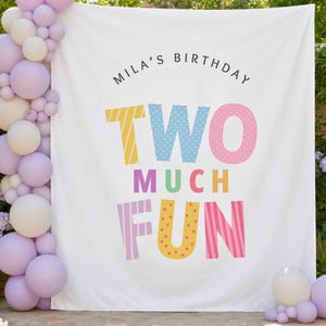 May include: A white fabric backdrop featuring the text "MILA'S BIRTHDAY" in black and "TWO MUCH FUN" in large, colorful, patterned letters. A vertical garland of light purple and white balloons is attached to the left side of the backdrop.