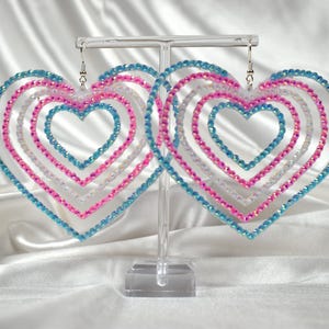 May include: A pair of heart-shaped earrings. Each earring features multiple nested heart outlines in blue, pink, and white, adorned with sparkling beads. The earrings are displayed on a clear stand against a white satin background.