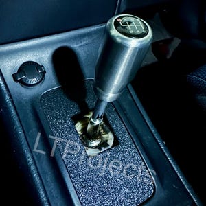 May include: A close-up of a silver gear shift knob with a black and white gear pattern on top. The knob is installed in a black car interior with a black textured surface.