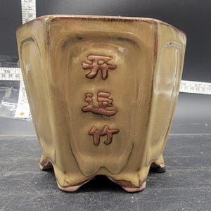 May include: A hexagonal ceramic planter in shades of tan and brown. The planter has a glossy finish and features embossed Chinese characters. The base has small feet. A ruler is visible in the background.