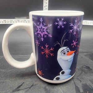 Puede incluir: Una taza de cer&aacute;mica blanca con fondo azul y una imagen colorida de Olaf de la pel&iacute;cula Frozen. La taza tiene un patr&oacute;n de copo de nieve y las palabras "Let It Go" est&aacute;n impresas en el lateral.