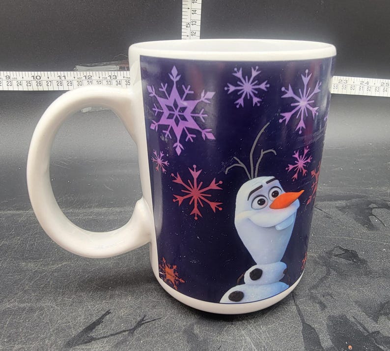 Puede incluir: Una taza de cer&aacute;mica blanca con fondo azul y una imagen colorida de Olaf de la pel&iacute;cula Frozen. La taza tiene un patr&oacute;n de copo de nieve y las palabras "Let It Go" est&aacute;n impresas en el lateral.