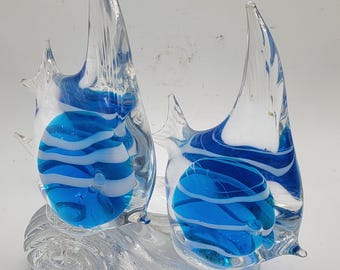 May include: A glass sculpture of two blue and white fish on a wave base. The fish are clear glass with blue and white swirls inside.