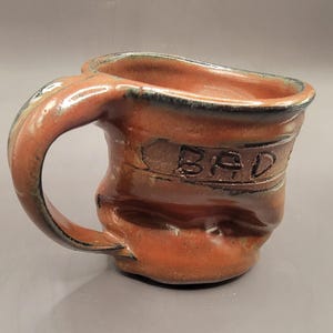 Puede incluir: Una taza de cerámica hecha a mano con un esmalte marrón y una forma ligeramente deformada. La taza tiene un asa y la palabra "BAD" está tallada en el lateral.