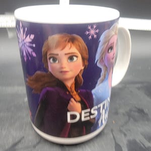 Puede incluir: Una taza de cer&aacute;mica blanca con fondo morado que presenta im&aacute;genes de Anna y Elsa de la pel&iacute;cula Frozen II. El texto "DESTINY" est&aacute; impreso en la taza.