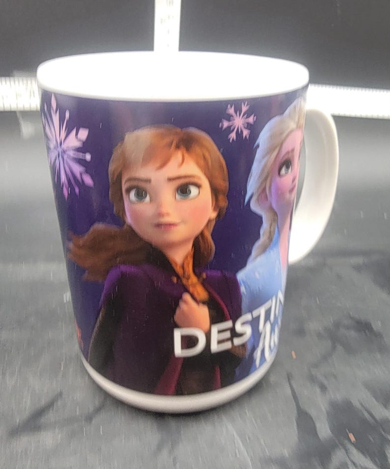 Puede incluir: Una taza de cer&aacute;mica blanca con fondo morado que presenta im&aacute;genes de Anna y Elsa de la pel&iacute;cula Frozen II. El texto "DESTINY" est&aacute; impreso en la taza.
