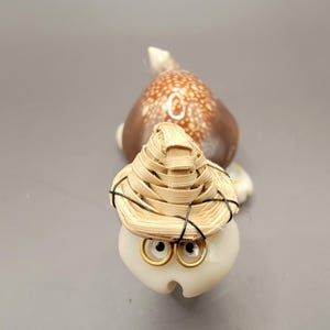 May include: A small, white seashell turtle figurine with a straw hat and gold-rimmed eyes. The turtle is wearing a straw hat with a black ribbon.