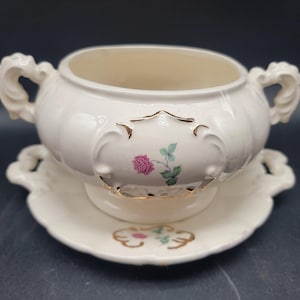 May include: A white porcelain soup tureen with a gold trim and floral design. The tureen has a matching saucer with a scalloped edge.
