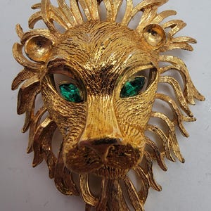 May include: A gold-toned lion brooch with green rhinestone eyes. The lion's mane is detailed with textured lines.