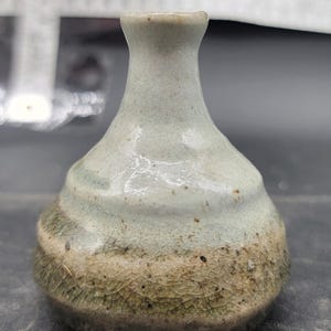 Può includere: Un piccolo vaso in ceramica fatto a mano con collo stretto e corpo arrotondato. Il vaso presenta una combinazione di colori a due toni, con una parte superiore verde chiaro e una sezione inferiore marrone strutturata. La superficie ha una finitura lucida.