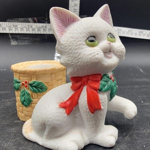 Op de afbeelding: Witte keramische kattenfiguur met een rode strik en hulstbladeren om zijn nek. De kat zit naast een kleine rieten mand met hulstbladeren en rode bessen.