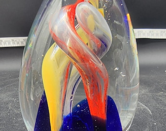 May include: A clear glass paperweight with a blue, yellow, and red swirl inside. The swirl is encased in clear glass and has a blue base.