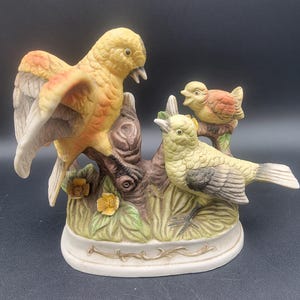 May include: A porcelain figurine of three yellow birds perched on a tree branch. The birds are painted in shades of yellow, orange, and brown. The figurine is set on a white base with a gold trim.