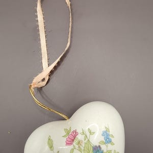 May include: A white porcelain heart-shaped ornament with a floral pattern in shades of blue, pink, and green. The ornament has a gold string attached to a cream-colored ribbon.