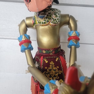 Puede incluir: Un títere de madera con la cara pintada y un elaborado disfraz. El títere sostiene un arma en su mano derecha. El títere está pintado en dorado, rojo, azul y verde.