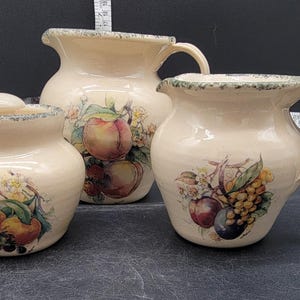 Könnte beinhalten: Set aus drei Keramik-Küchenbehältern mit cremefarbenem Hintergrund und Blumendesigns mit Früchten. Die Behälter haben einen grünen Rand und sind mit Pfirsichen, Pflaumen und Trauben verziert.